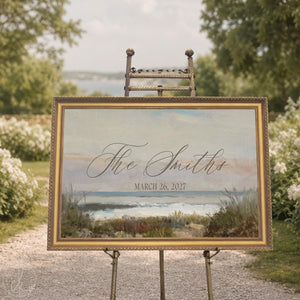 Beach wedding decor sign with a vintage seascape canvas in an ornate gold frame on a brass easel, script last name and date.