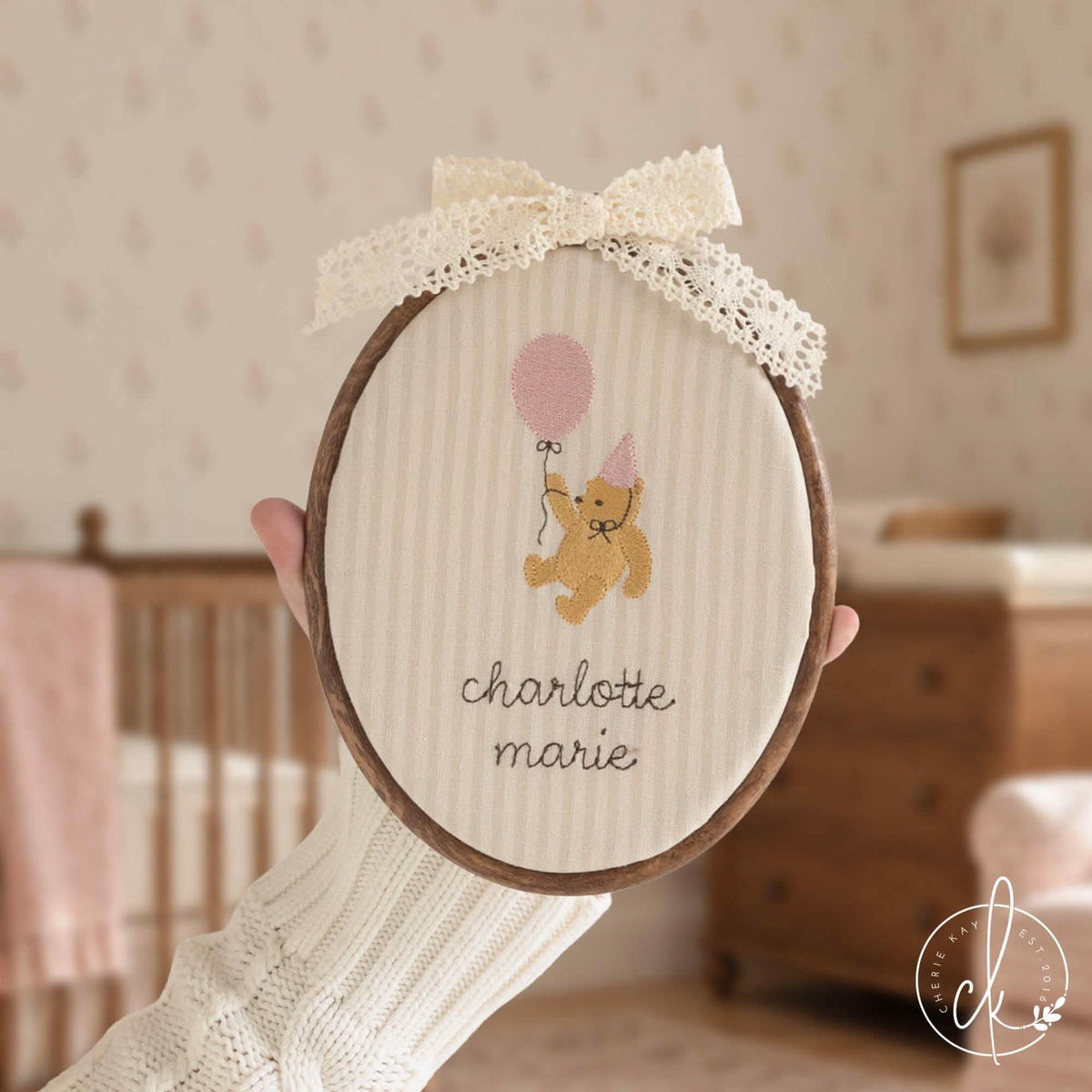 Winnie the Pooh gift name hoop with a bear holding a pink balloon over a cursive name, held up in a girl nursery