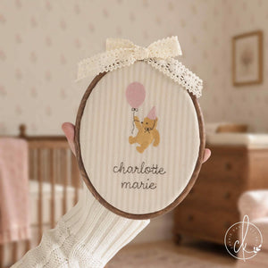 Winnie the Pooh gift name hoop with a bear holding a pink balloon over a cursive name, held up in a girl nursery