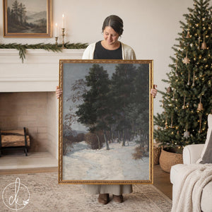 Winter home decor vertical painting of dark evergreens in snow in an ornate gold frame, held up in a living room