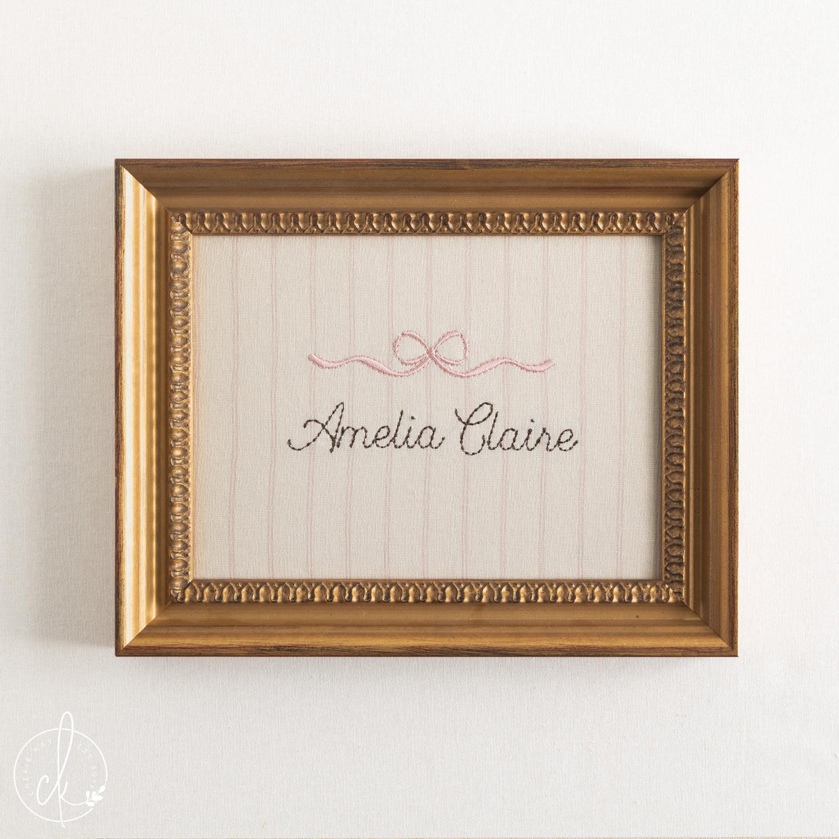 Girl nursery decor name sign, pink embroidered bow over a cursive name on striped cream fabric in an ornate gold frame