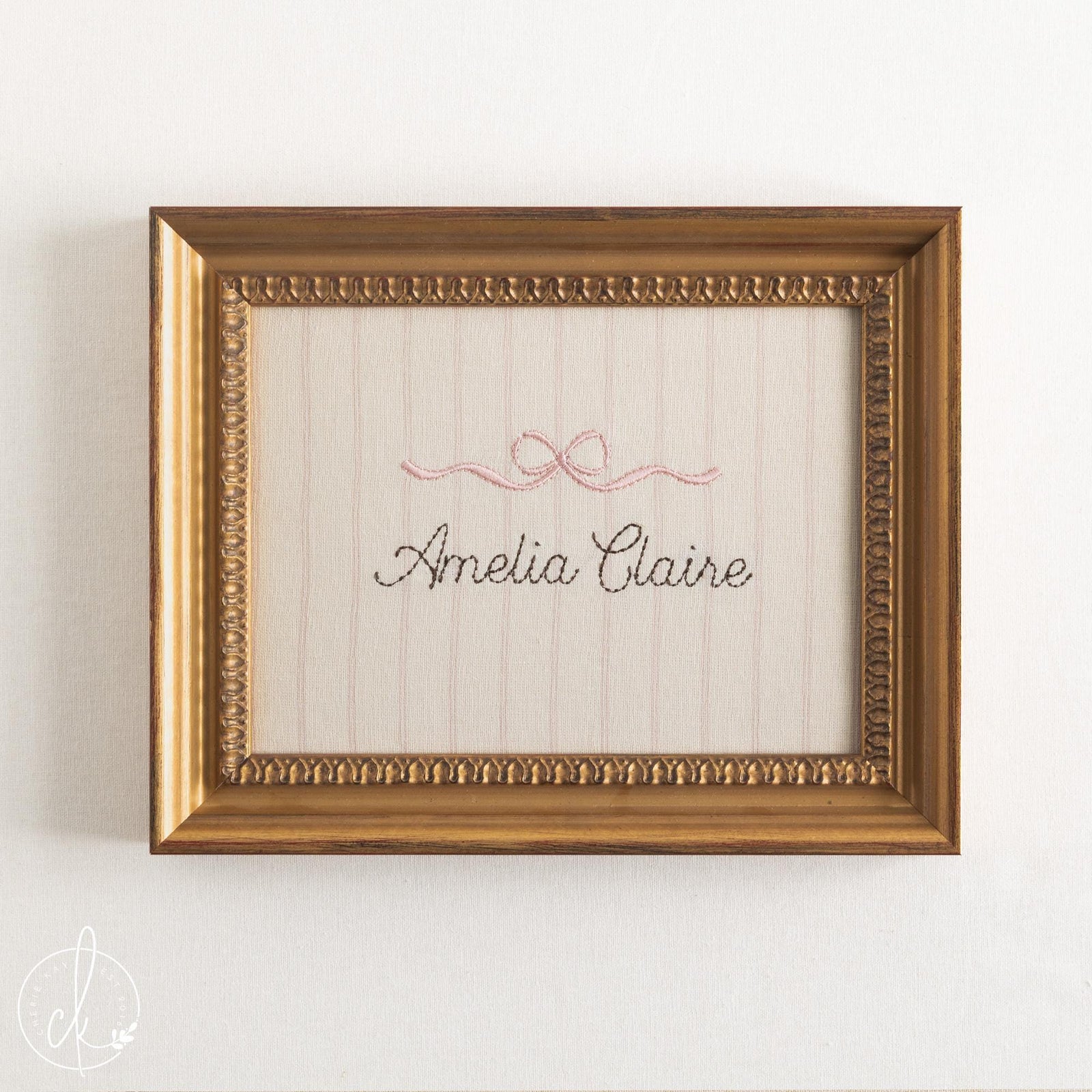 Girl nursery decor name sign, pink embroidered bow over a cursive name on striped cream fabric in an ornate gold frame