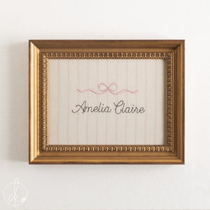Girl nursery decor name sign, pink embroidered bow over a cursive name on striped cream fabric in an ornate gold frame