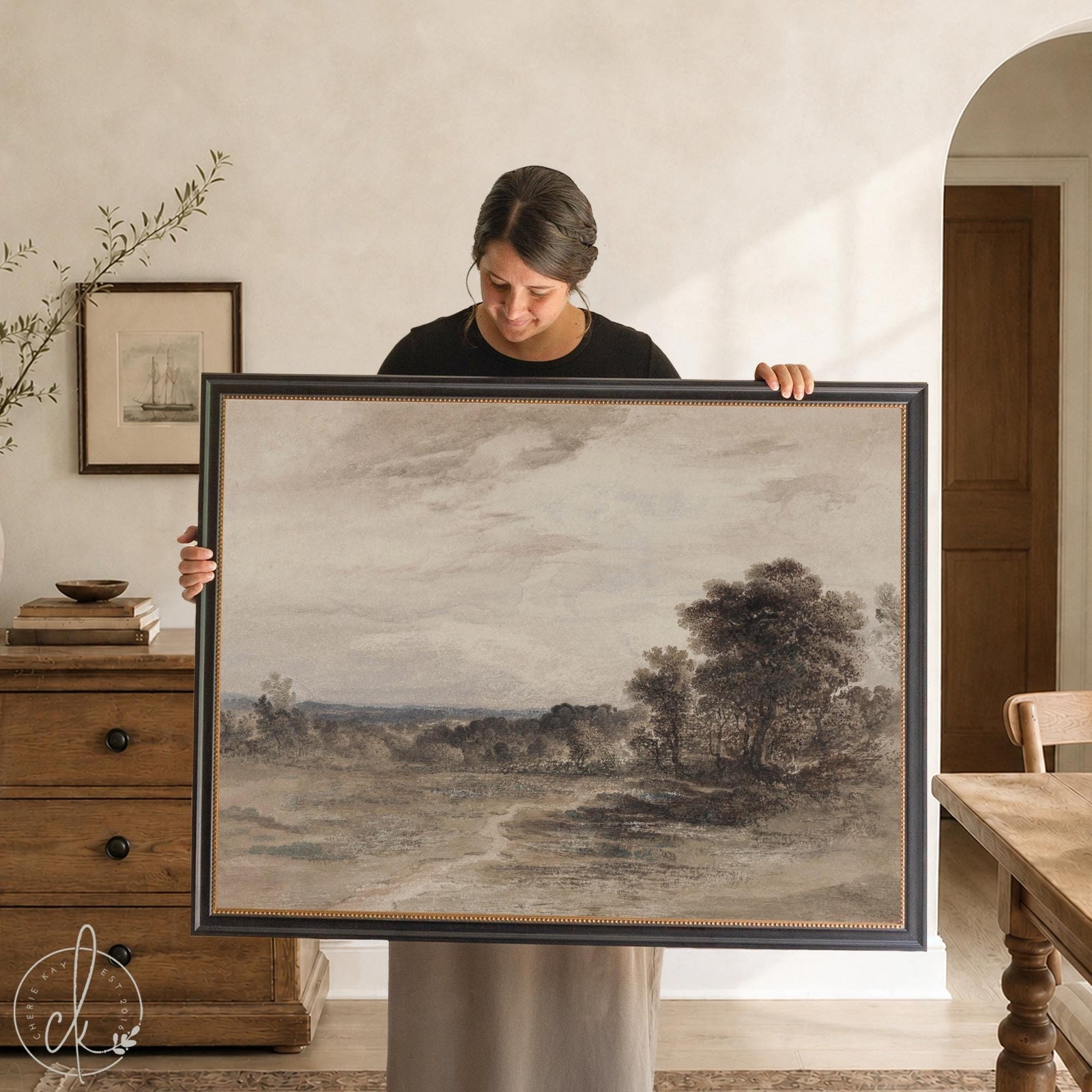 Moody art print of a countryside landscape under a cloudy sky in a black and gold frame, held up in a dining room