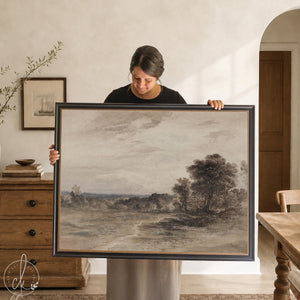 Moody art print of a countryside landscape under a cloudy sky in a black and gold frame, held up in a dining room