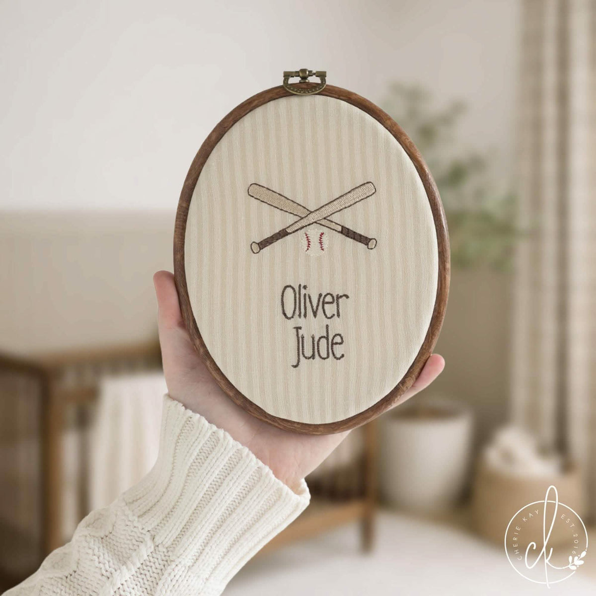 Baseball nursery decor name hoop with crossed bats and a baseball on cream striped fabric, held up in a nursery