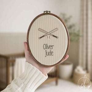 Baseball nursery decor name hoop with crossed bats and a baseball on cream striped fabric, held up in a nursery