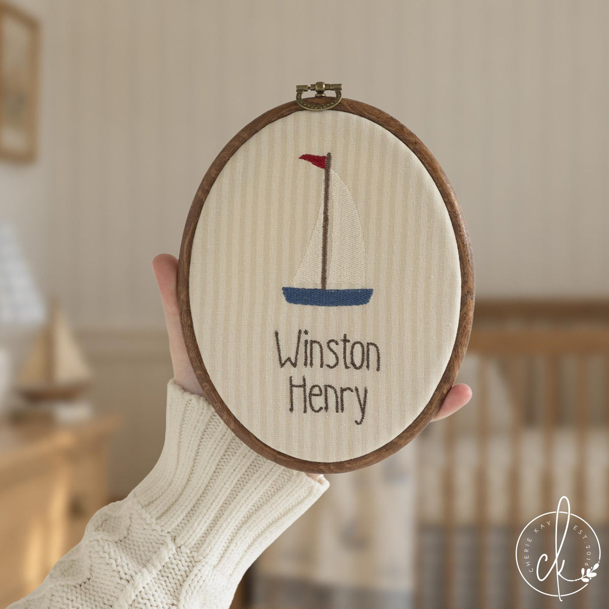 Nautical nursery decor hoop with a navy and cream sailboat and a baby name in brown letters, held up in a nursery