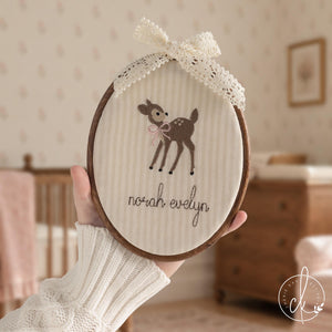 Woodland nursery decor hoop with a brown fawn in a pink bow and a baby name in brown cursive, held up in a nursery