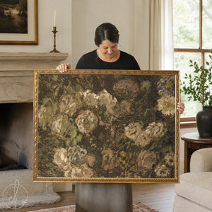 Monet flowers painting in an ornate gold frame, cream blooms on a dark brown background, held up in a living room