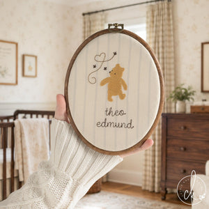 Pooh bear decor hoop with a honey gold bear, three bees, a heart trail and a baby name in brown script, held up in a nursery.