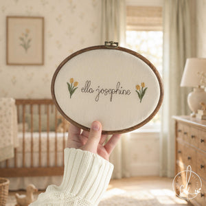 Newborn keepsake hoop with yellow tulips and a baby name in brown cursive on cream fabric, held up in a nursery