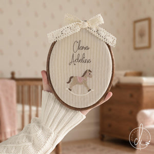 Unique baby gifts hoop with a pink rocking horse, a baby name in brown script and a cream lace bow, held up in a nursery