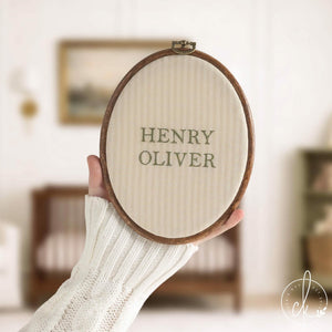 Baby boy gift name hoop with a sage green serif name on cream striped fabric, held up in a nursery