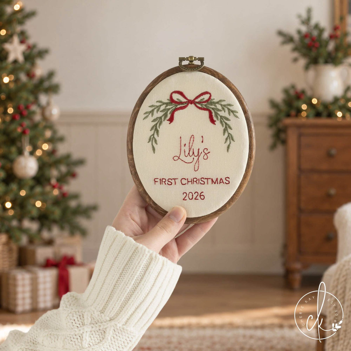 Newborn Christmas gift name hoop with a red bow, green garland and red script name on cream fabric, held up in a living room