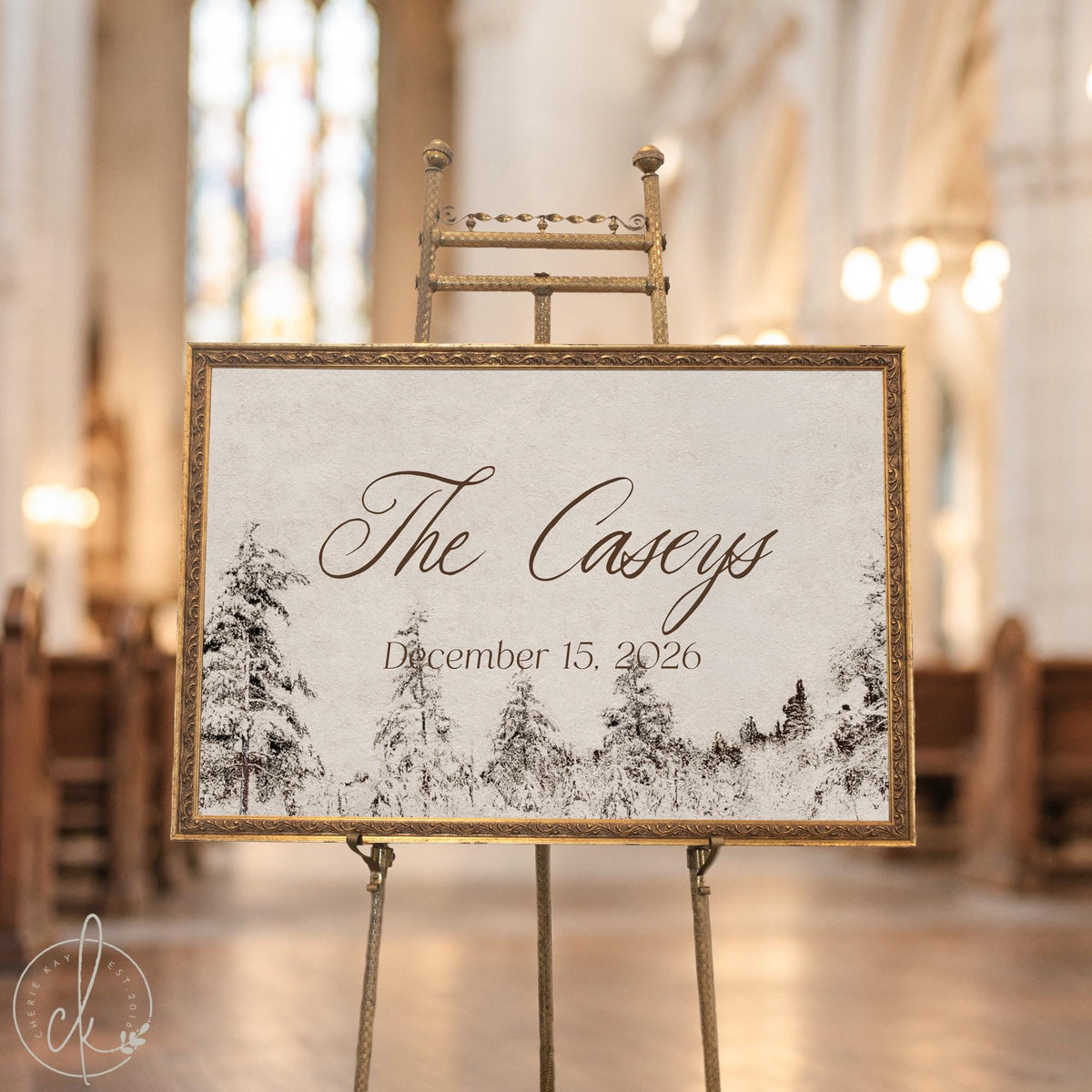 Winter wedding decor sign with a last name and date over snowy pine trees in an ornate gold frame on an easel