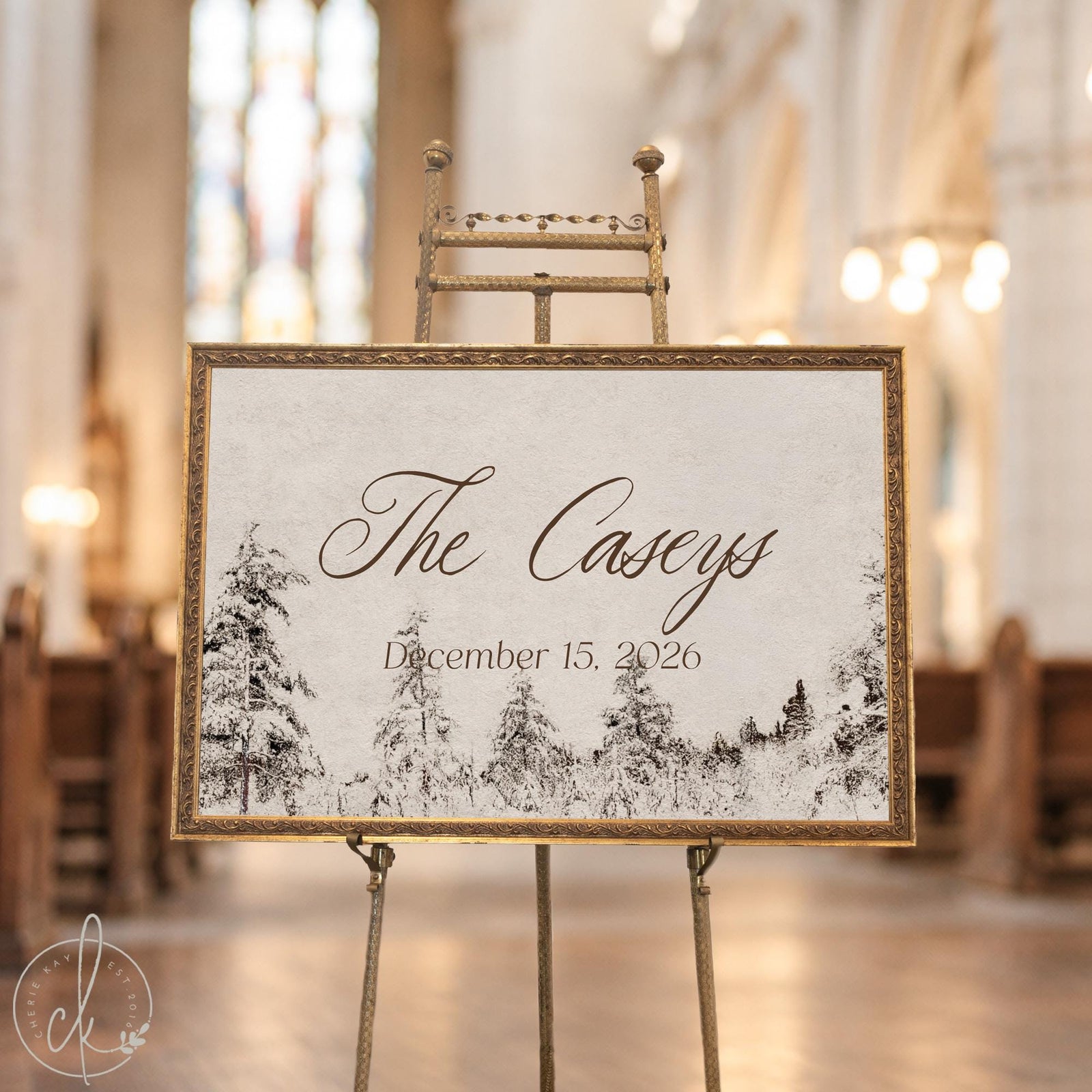 Winter wedding decor sign with a last name and date over snowy pine trees in an ornate gold frame on an easel