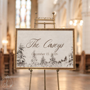 Winter wedding decor sign with a last name and date over snowy pine trees in an ornate gold frame on an easel