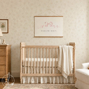 Personalized wall hanging with a pink watercolor bow and baby name on canvas, hung above a wood crib in a nursery