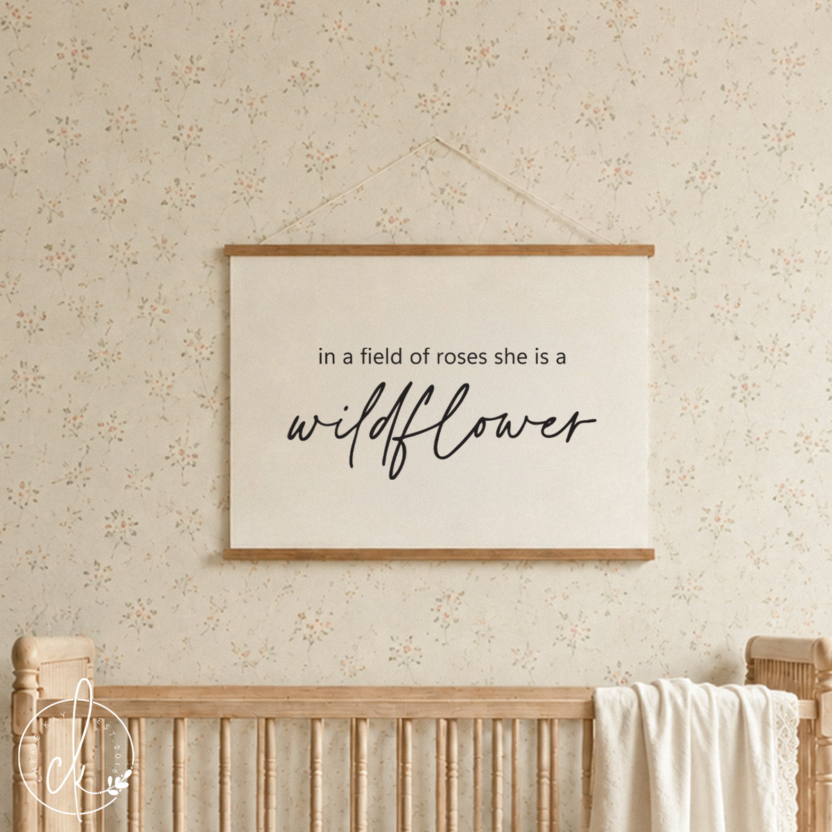  A rectangular wall hanging featuring a light-colored fabric canvas held by two horizontal wooden bars. The text "in a field of roses she is a wildflower" is printed in black, with the word wildflower in a cursive script font.