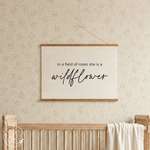  A rectangular wall hanging featuring a light-colored fabric canvas held by two horizontal wooden bars. The text "in a field of roses she is a wildflower" is printed in black, with the word wildflower in a cursive script font.