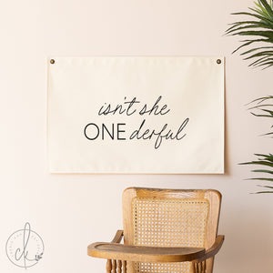 Decorative sign with 'isn't she ONEderful' text above a wicker chair on a beige wall.