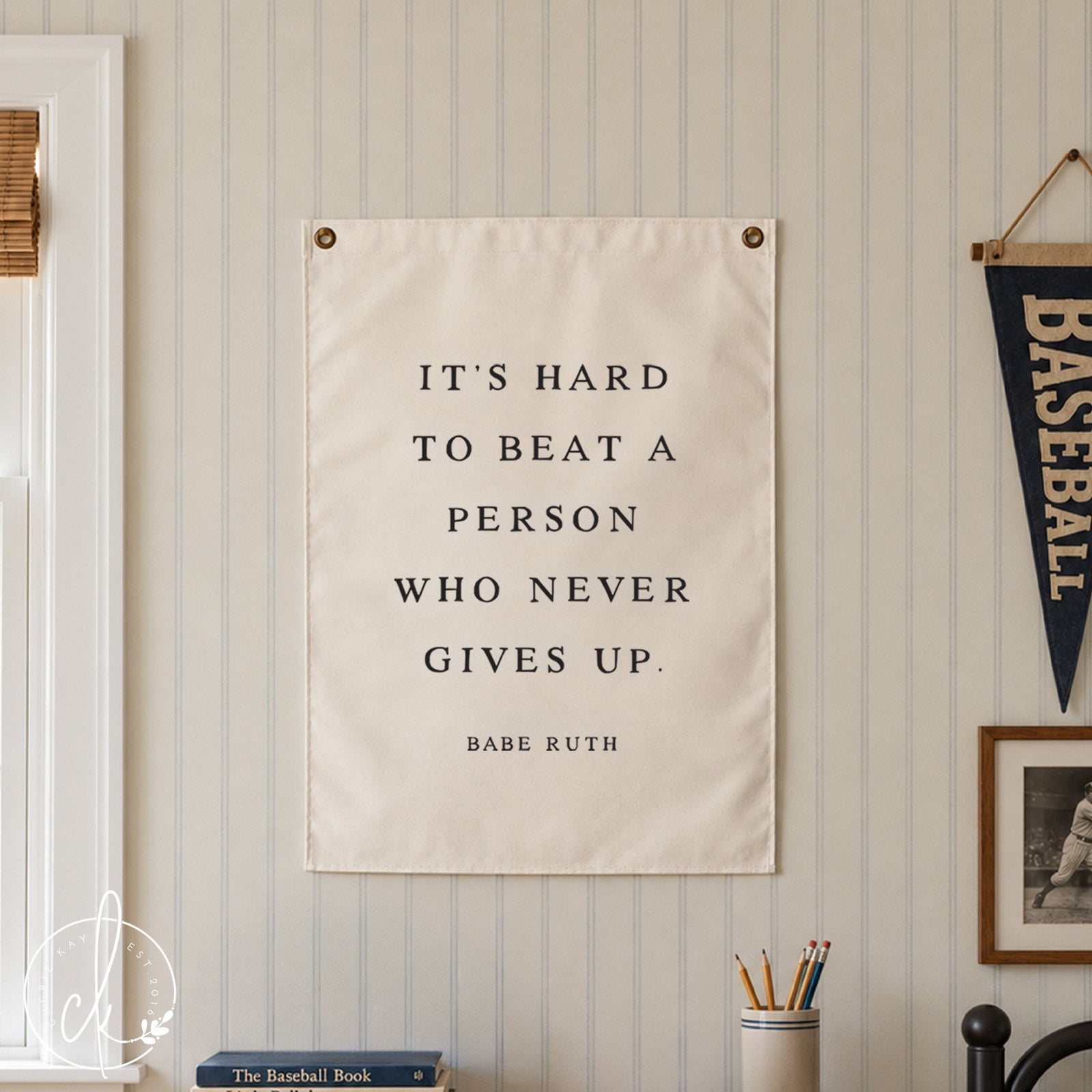 A white rectangular wall hanging featuring the black text: IT'S HARD TO BEAT A PERSON WHO NEVER GIVES UP. BABE RUTH. The sign is mounted with two metal grommets at the top corners and displays a motivational quote in a serif font.