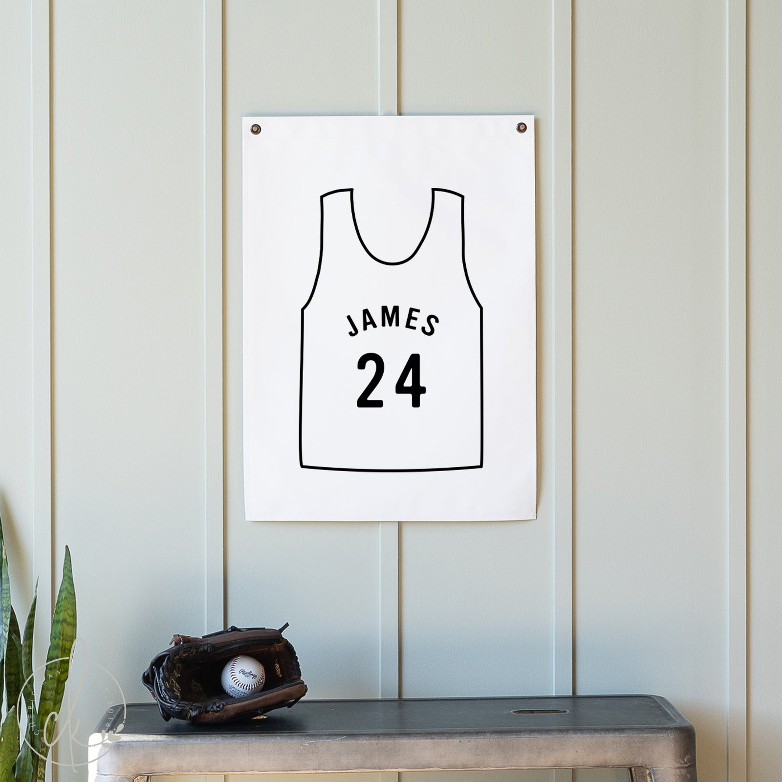 White wall with a framed black and white illustration of a basketball jersey with 'JAMES 24' on it, next to a baseball glove and ball on a wooden surface.