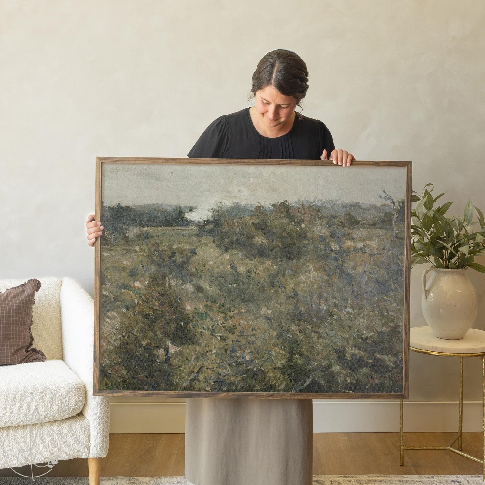 Large landscape wall art featuring abstract countryside scene with rolling hills and muted earth tones, displayed by woman in modern living room setting with neutral decor and natural wood frame