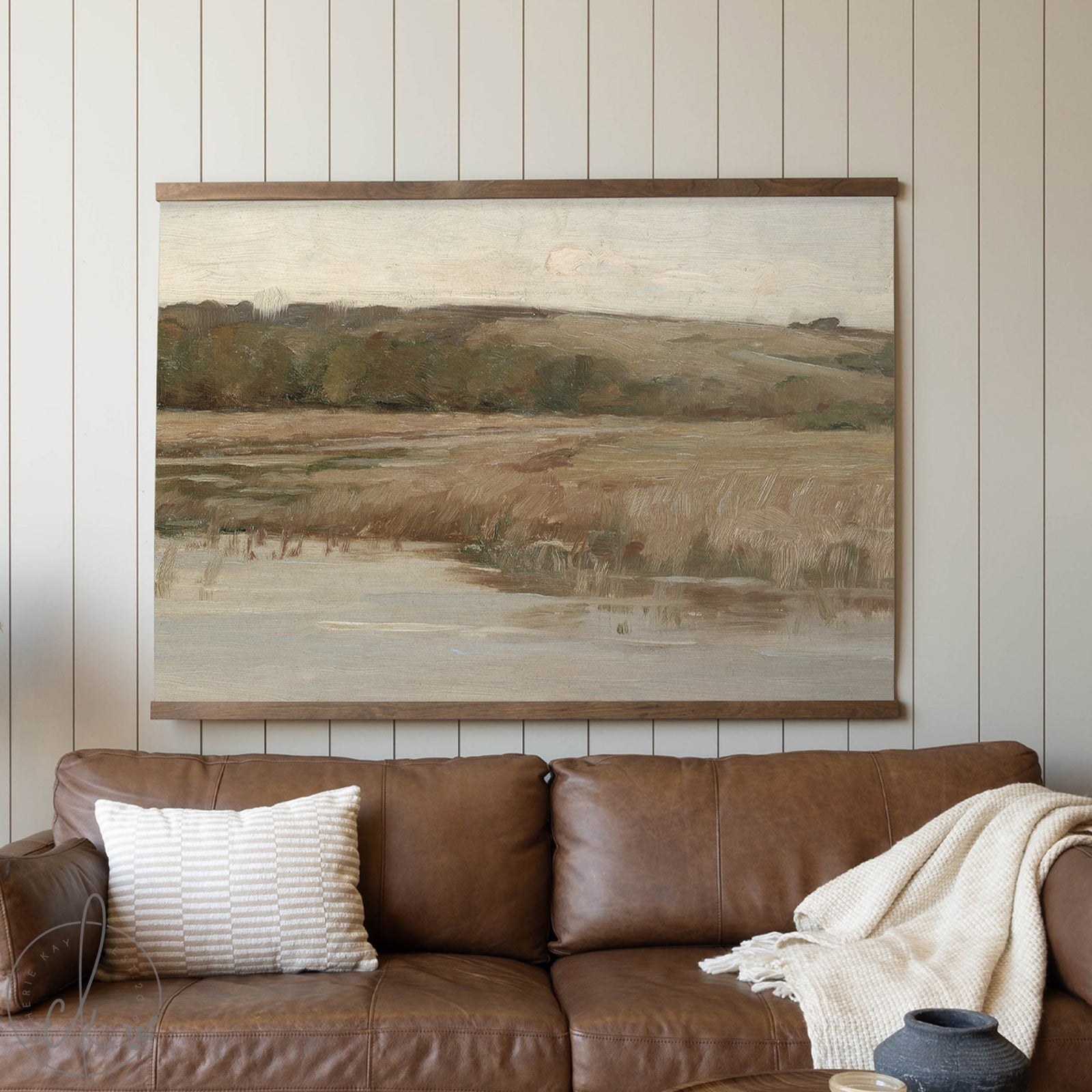 Brown leather sofa with a blanket and pillow in front of a landscape painting on a wooden wall.