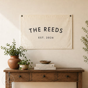 Personalized canvas banner hanging on a white brick wall, featuring the text 'The Halls, Est. 2024.' The sign is displayed near a black side table with a small potted plant and candle, adding a modern rustic touch to the entryway decor.