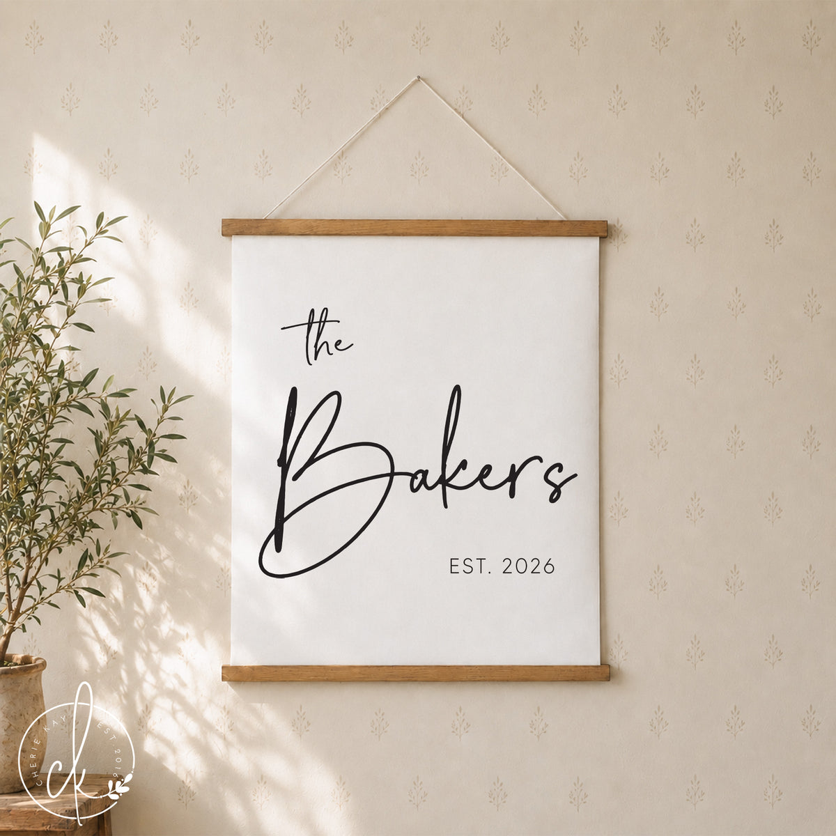 A white canvas print with black text that reads "the Williams EST. 2010" hanging on a dark gray wall. The print is framed with a natural wood frame and is hanging from a white string.