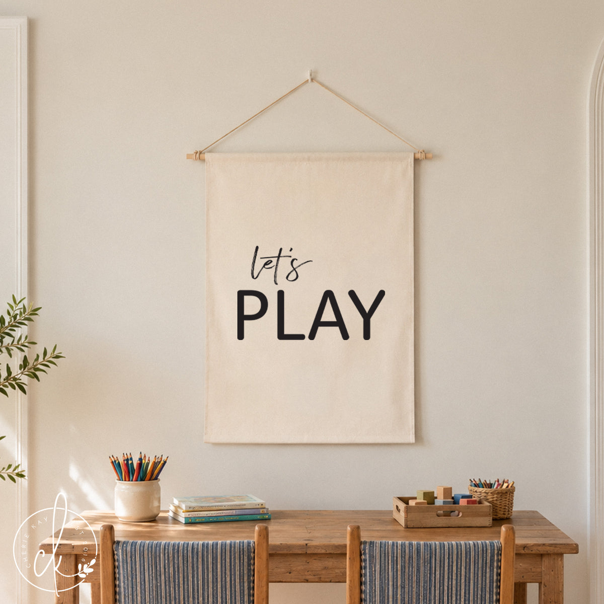 A white fabric wall hanging with the black text "let's PLAY" suspended by a string. The banner is displayed above a light wood table with two matching chairs