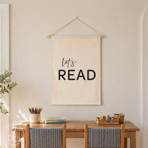A white fabric wall hanging with the black text "let's READ" suspended by a wooden dowel and string. The banner is displayed above a small wooden table with two matching chairs
