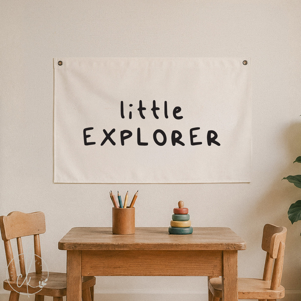An off-white fabric banner with the words 'little EXPLORER' printed in a black handwritten font, hanging on a beige wall above a wooden children's table and chairs.