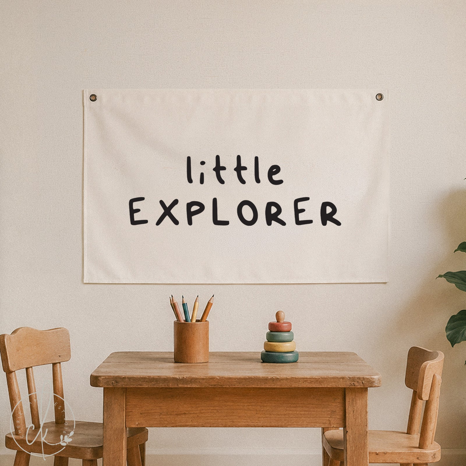 An off-white fabric banner with the words 'little EXPLORER' printed in a black handwritten font, hanging on a beige wall above a wooden children's table and chairs.