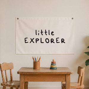 An off-white fabric banner with the words 'little EXPLORER' printed in a black handwritten font, hanging on a beige wall above a wooden children's table and chairs.