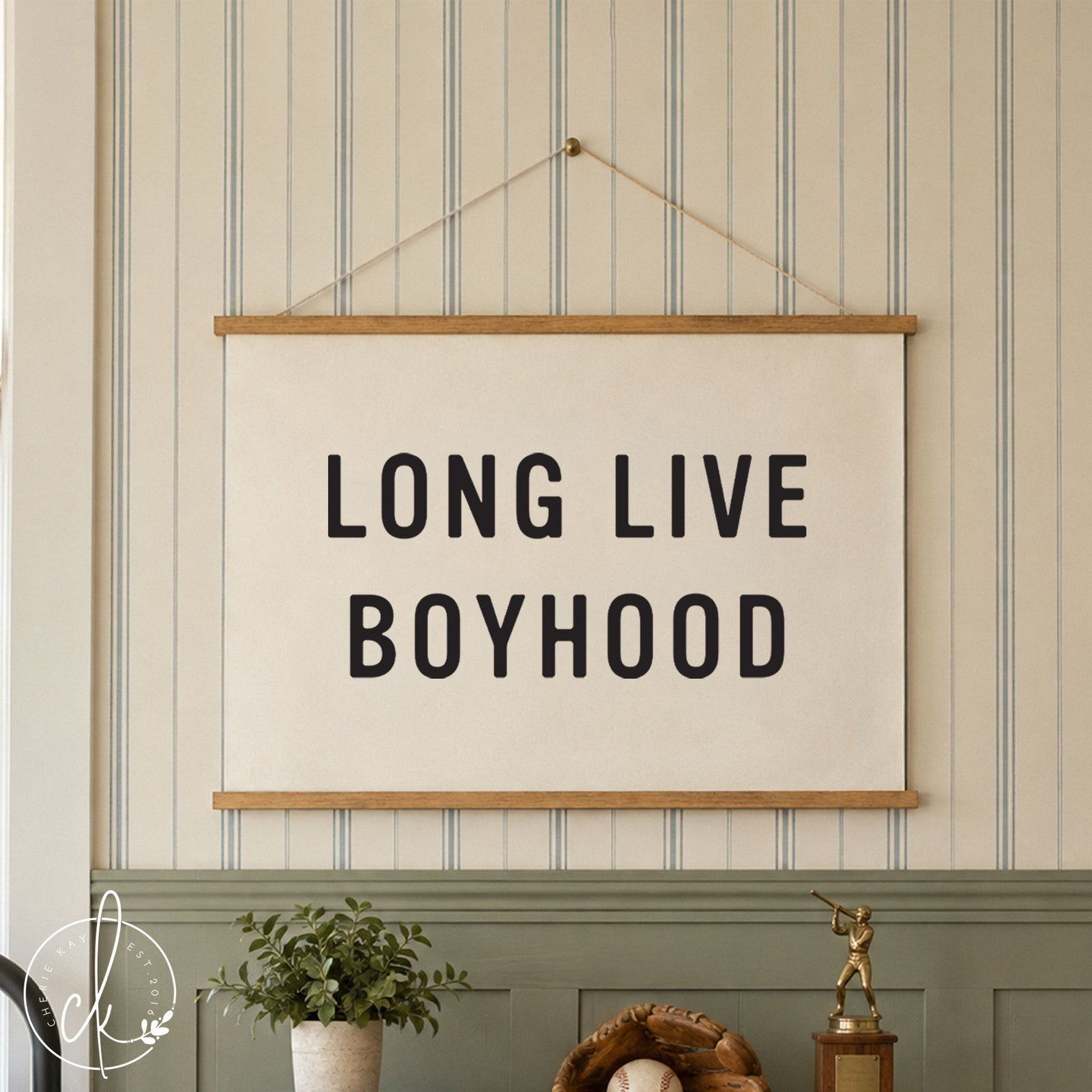 A white wall hanging with the black text LONG LIVE BOYHOOD, framed by two horizontal dark wood bars. The piece is suspended by a thin string, featuring a clean, minimalist design that displays bold, sans-serif typography.
