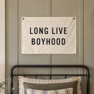 A rectangular white fabric wall banner featuring the black text LONG LIVE BOYHOOD in a bold sans-serif font. The banner is mounted with metal grommets in the top corners, displaying a clean, minimalist design for wall decor.