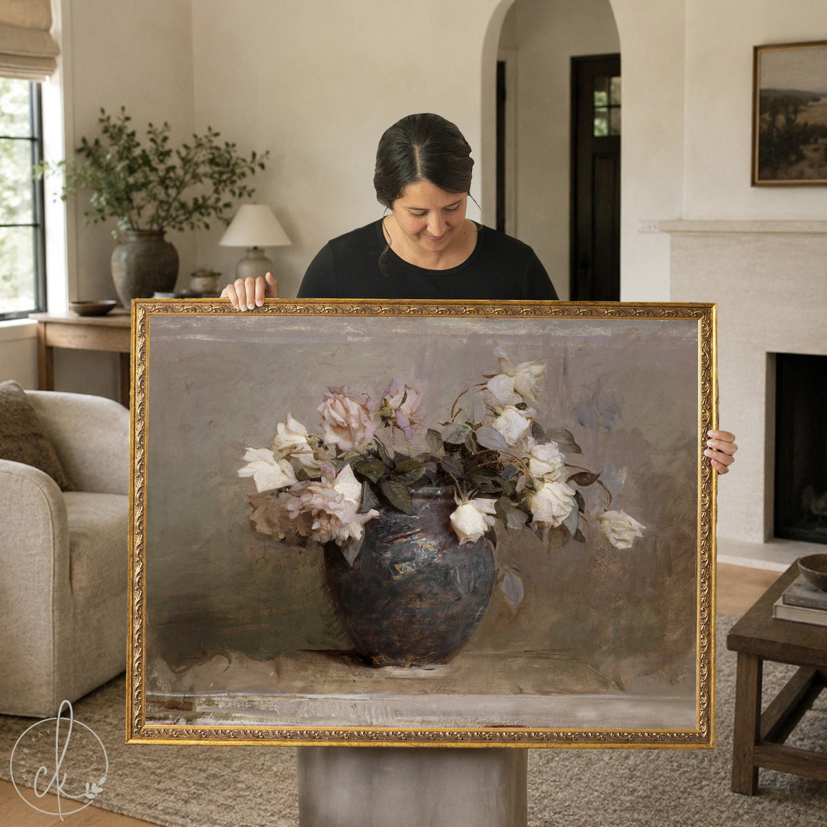 A woman holds a large framed vintage botanical art print featuring soft white and blush roses in a rustic blue ceramic vase. The floral still life has a muted, antique finish and is ideal for farmhouse or traditional home decor styles.