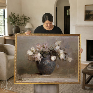 A woman holds a large framed vintage botanical art print featuring soft white and blush roses in a rustic blue ceramic vase. The floral still life has a muted, antique finish and is ideal for farmhouse or traditional home decor styles.