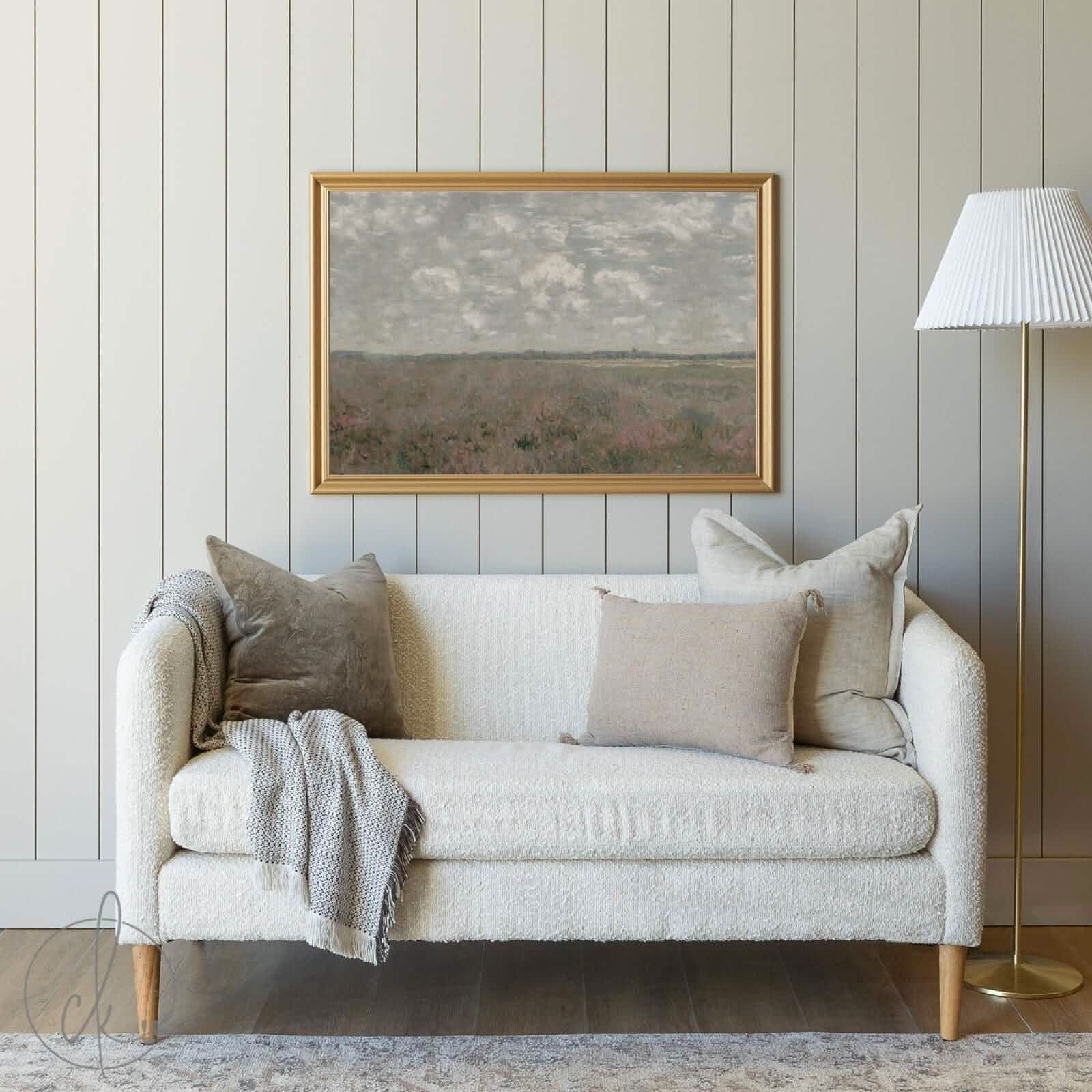 A cozy living room with a cream sofa, neutral pillows, a gray blanket, and a framed painting of a cloudy sky over a serene landscape on a muted paneled wall. A gold floor lamp with a white shade stands nearby.
