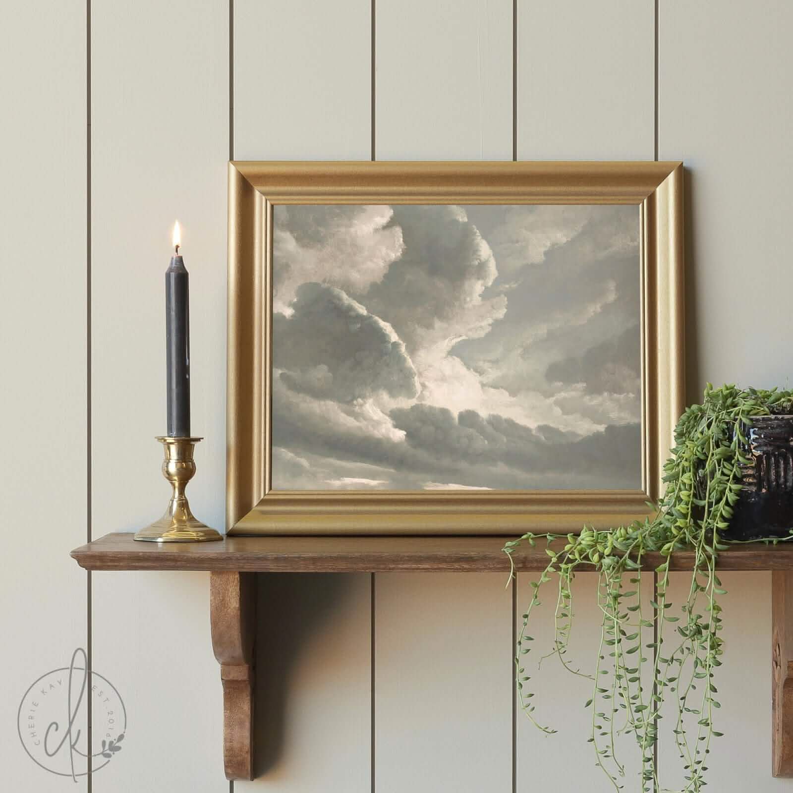 A framed painting of dramatic, cloudy skies with soft light breaking through, displayed on a wooden shelf alongside a lit black candle in a brass holder and a trailing green plant.