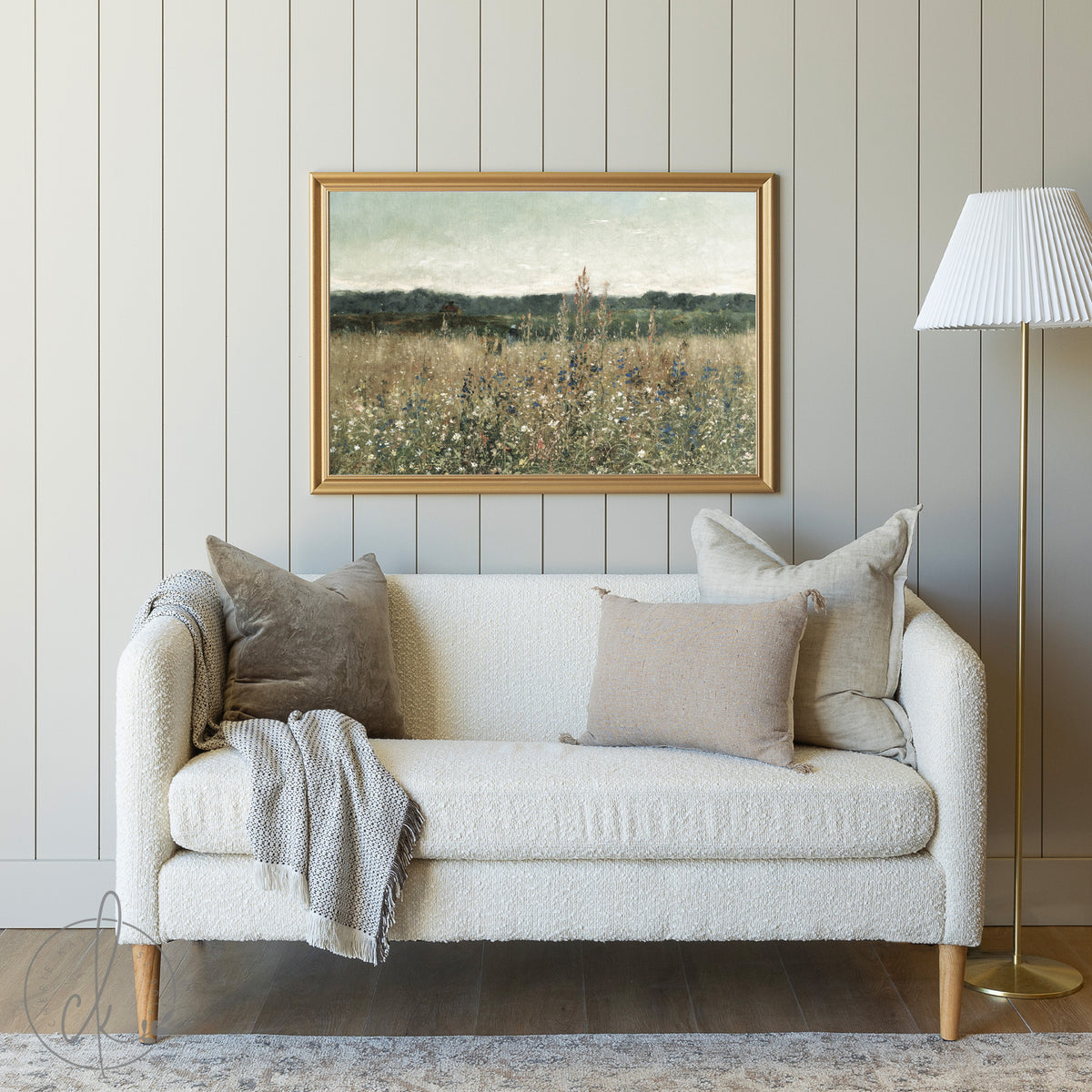 A cozy living room with a cream sofa, neutral pillows, a gray blanket, and a framed meadow painting on a muted paneled wall. A gold floor lamp with a white shade stands to the side.