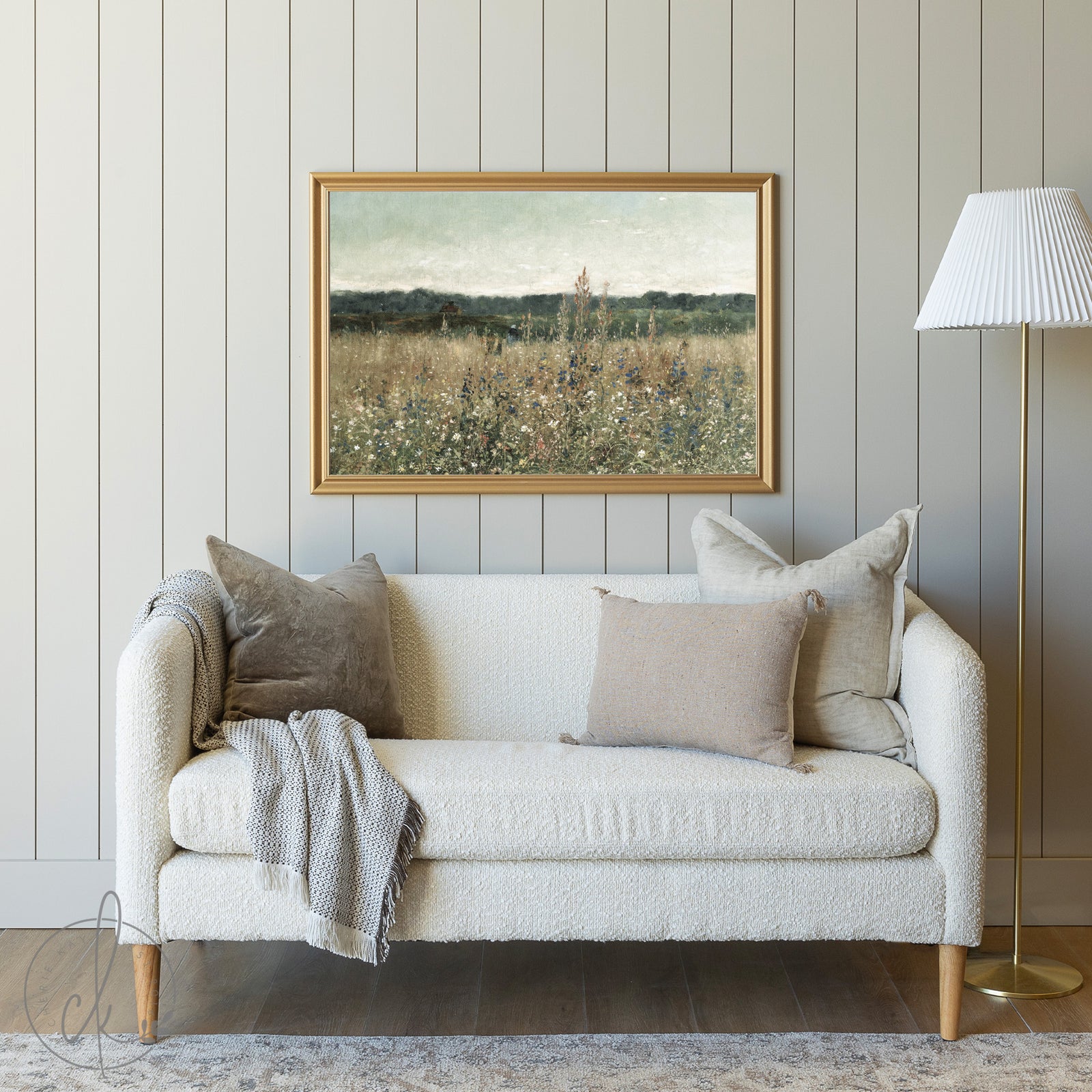 A cozy living room with a cream sofa, neutral pillows, a gray blanket, and a framed meadow painting on a muted paneled wall. A gold floor lamp with a white shade stands to the side.