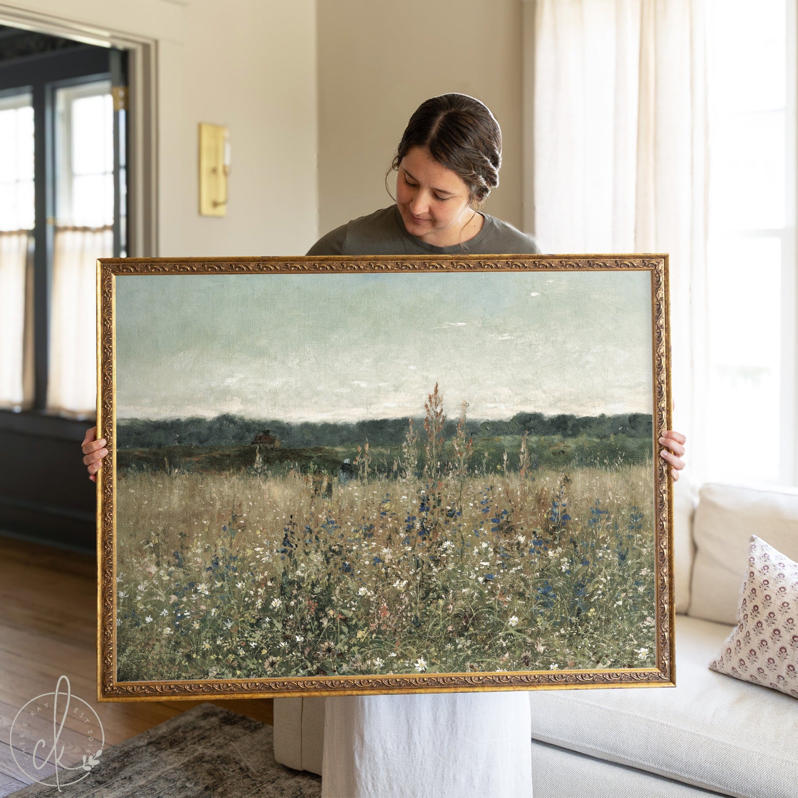 A woman holding a large framed botanical wall art painting featuring a serene wildflower meadow with tall grasses and distant tree-covered hills under a soft, overcast sky. The artwork is bordered by an ornate gold frame and is displayed in a warmly lit living room.