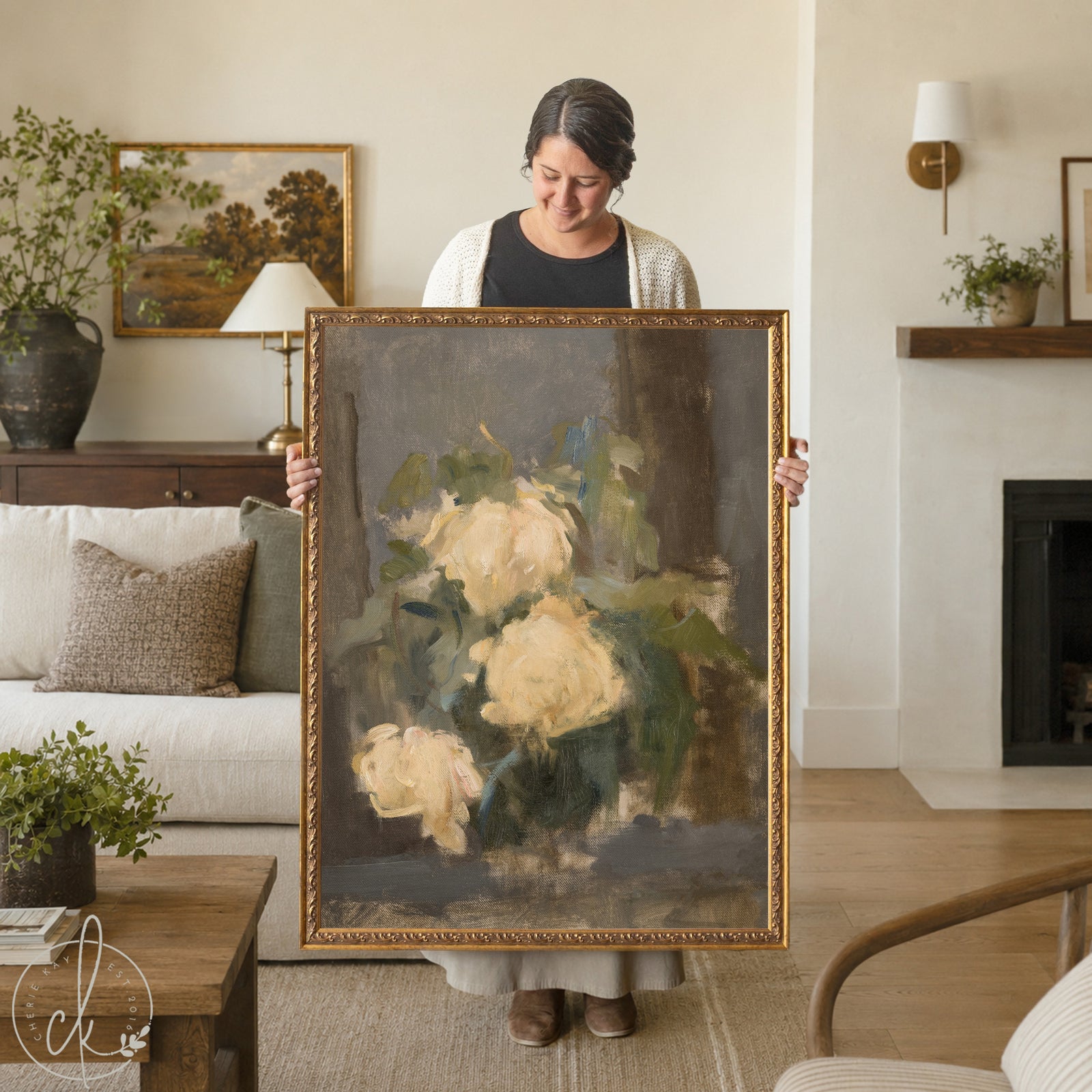 A rectangular oil painting in a gold ornate frame featuring a still life of cream-colored flowers with green foliage against a dark, moody gray background. The brushwork is visible and expressive, capturing the soft petals of the blooms.