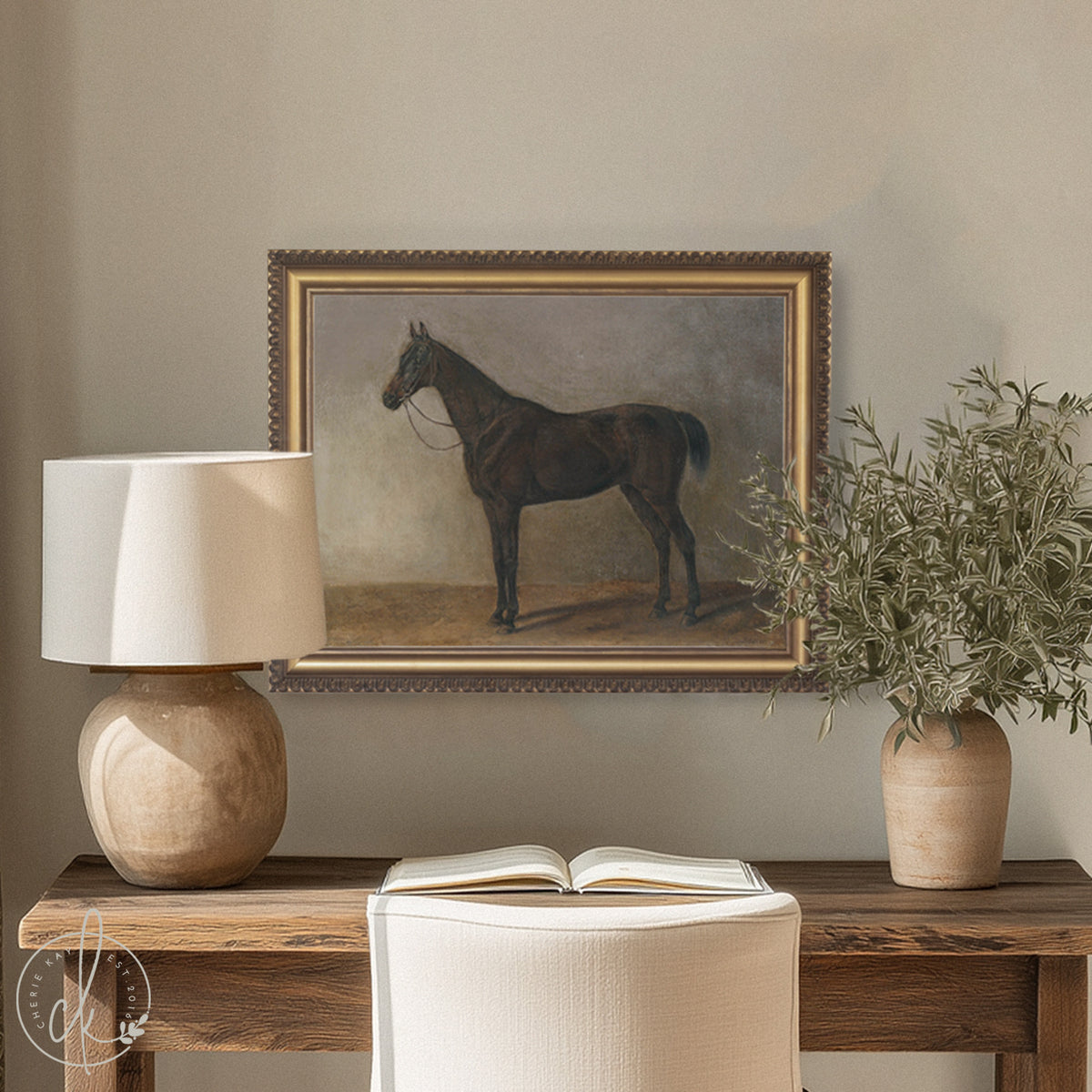 Moody antique horse print in an ornate gold frame above a rustic wood console with a lamp, open book, and olive branch vase.