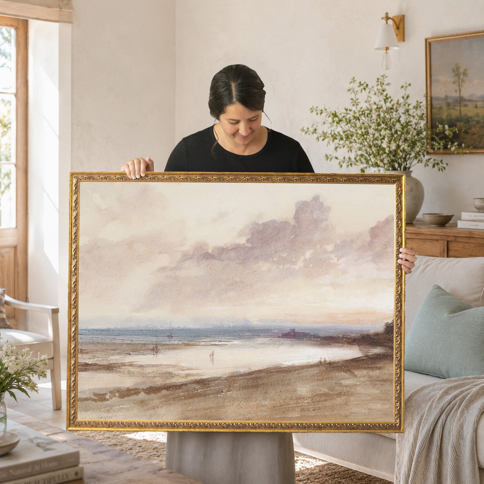 A rectangular landscape painting in a gold ornate frame depicting a coastal scene with a sandy beach, calm water, and a cloudy sky in muted brown, beige, and soft blue tones. The artwork features distant buildings and small figures.