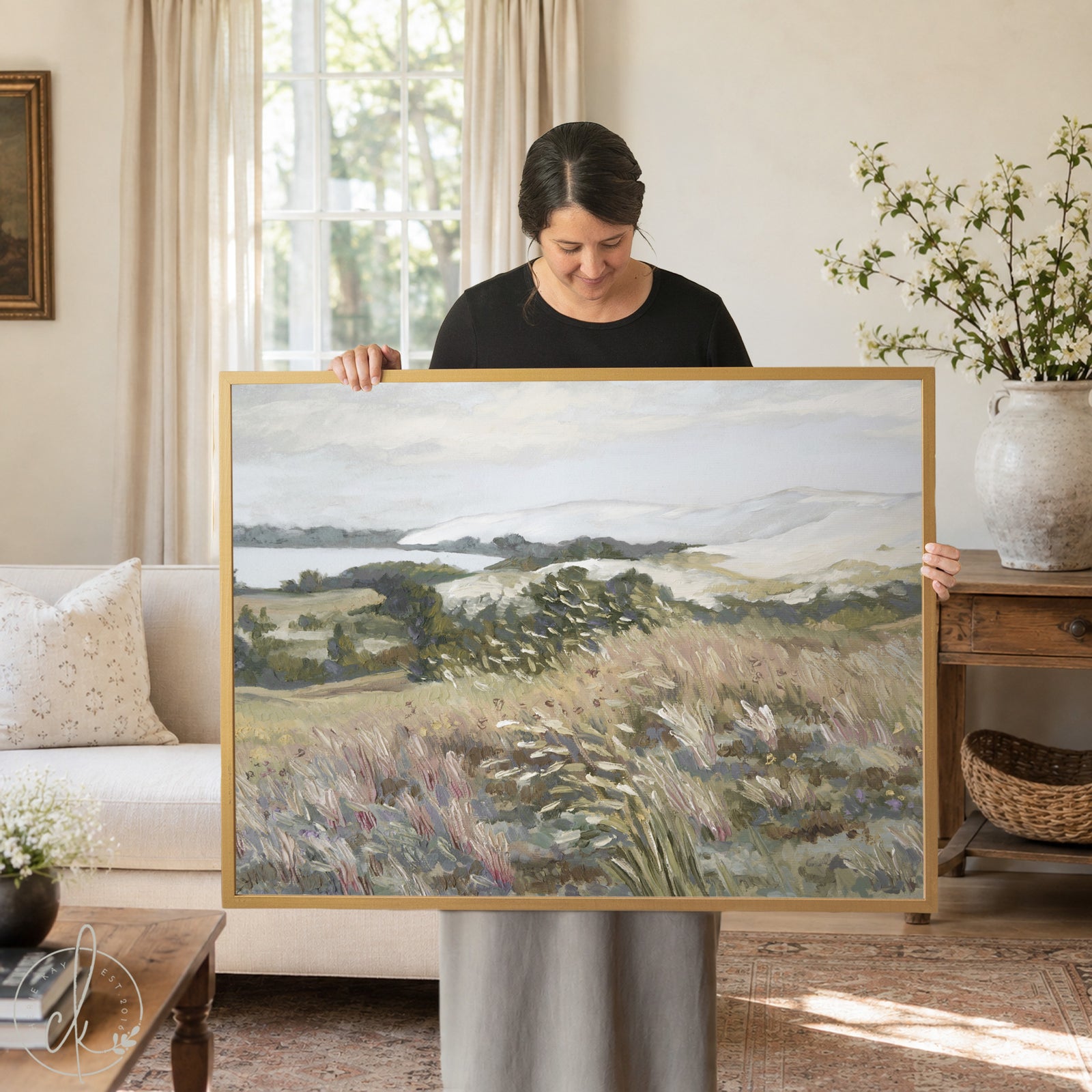 A framed landscape painting depicting a serene coastal scene. The artwork features a muted color palette of greens, browns, and blues, with a focus on grassy fields and distant hills. The frame is a warm, natural wood.